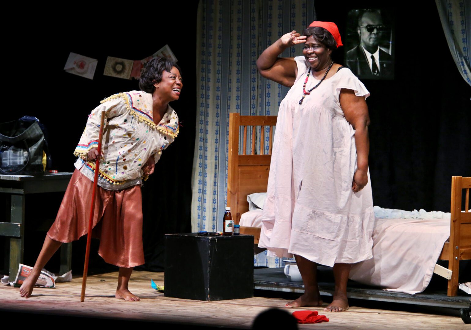 Two women on a stage in front of a bed.
