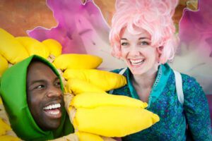 A man and woman holding bananas in front of a pink hair wig.